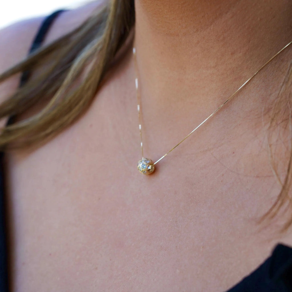 A woman elegantly wears the 14K Gold Bead Charm Pendant on a delicate gold chain. The diamond-cut finish and cubic zirconia sparkle as they catch the light, adding a touch of timeless sophistication to her outfit. The pendant rests gracefully on her collarbone, showcasing its minimalist yet luxurious design.