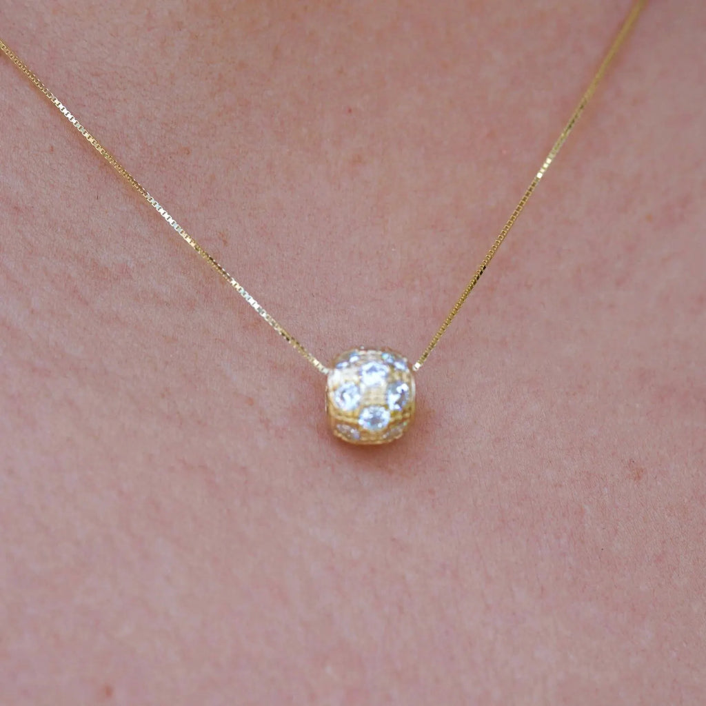 A woman elegantly wears the 14K Gold Bead Charm Pendant on a delicate gold chain. The diamond-cut finish and cubic zirconia sparkle as they catch the light, adding a touch of timeless sophistication to her outfit. The pendant rests gracefully on her collarbone, showcasing its minimalist yet luxurious design.