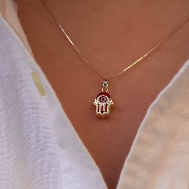 A model wears a 14K gold Hamsa pendant featuring a bold red Evil Eye, resting gracefully on the neckline. The vibrant red detail adds a unique touch of energy and charm to the protective talisman.