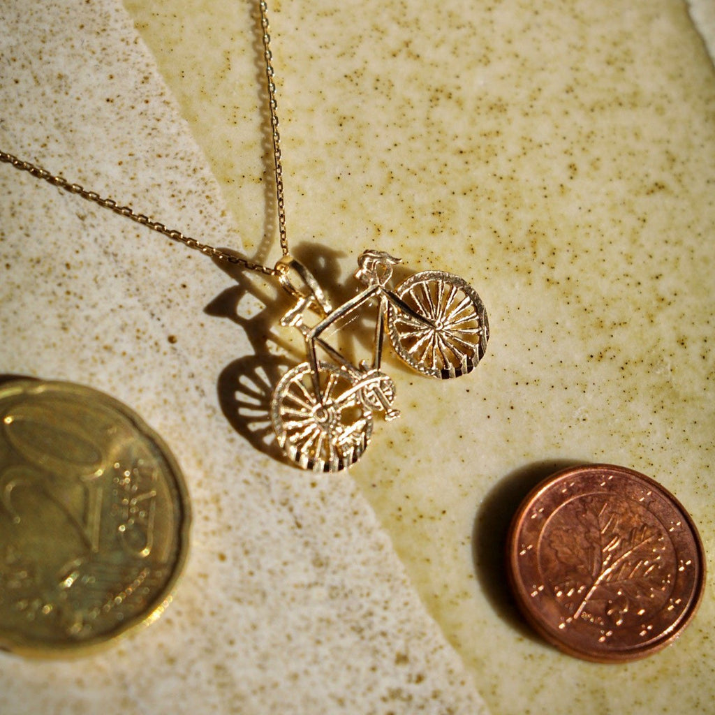 14K Solid Gold Bicycle Charm on a chain, placed next to a couple of coins for size comparison, captured from a slightly tilted angle to showcase its elegant craftsmanship and relative size.