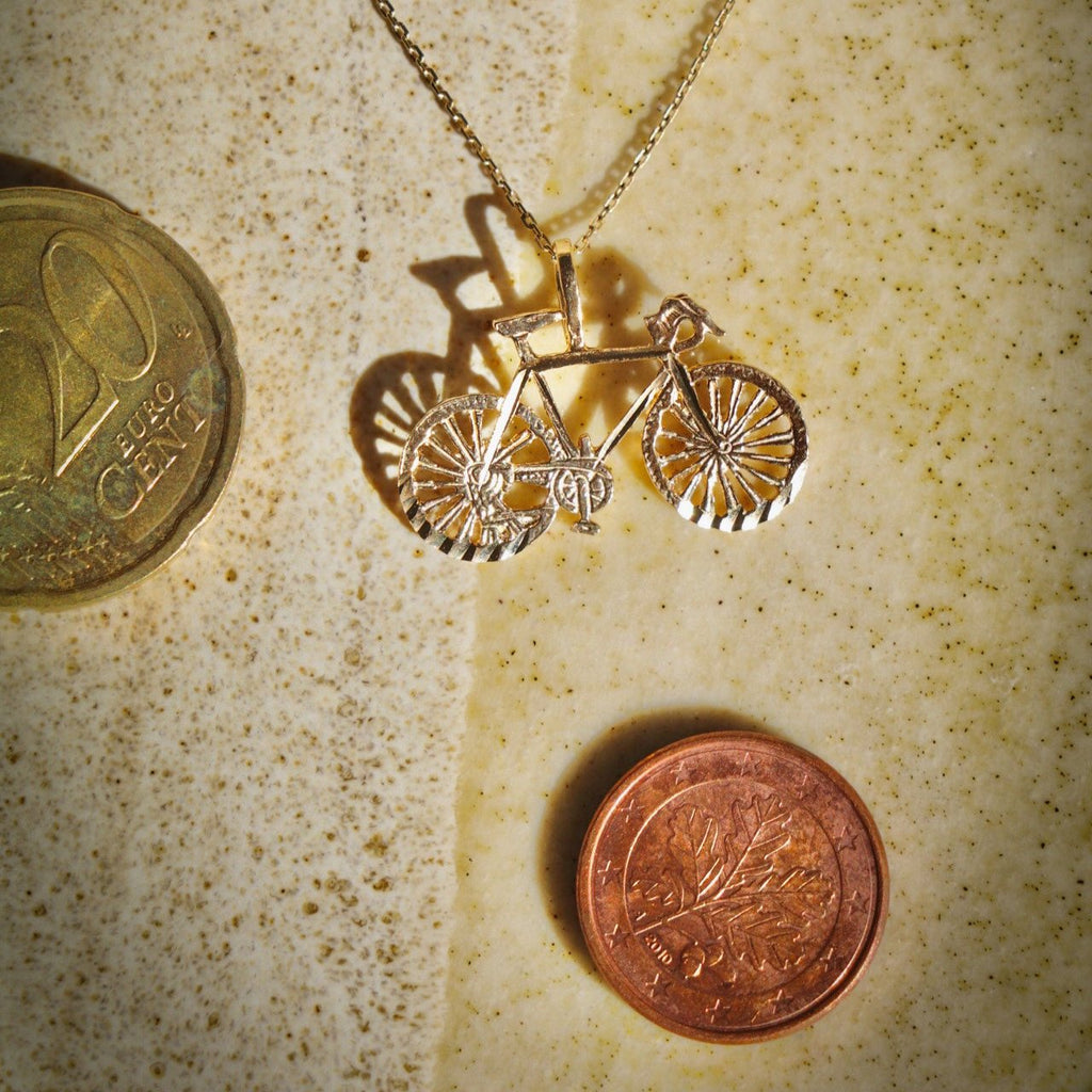 14K Solid Gold Bicycle Charm displayed on a chain next to a couple of coins for size comparison, shown in an upright position to highlight its dimensions and polished design.
