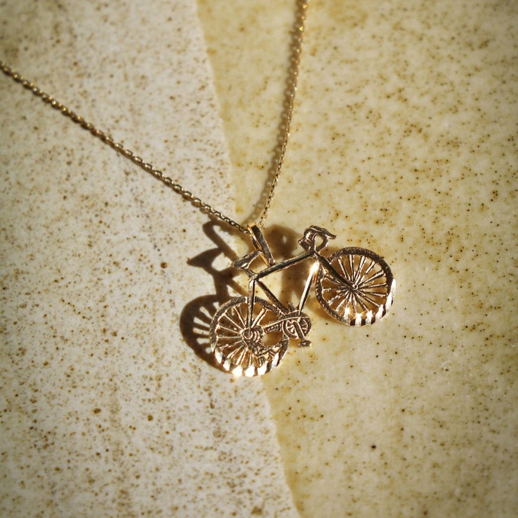 14K Solid Gold Bicycle Charm attached to a chain, resting on a flat surface with a slight tilt to showcase its polished finish and intricate design.