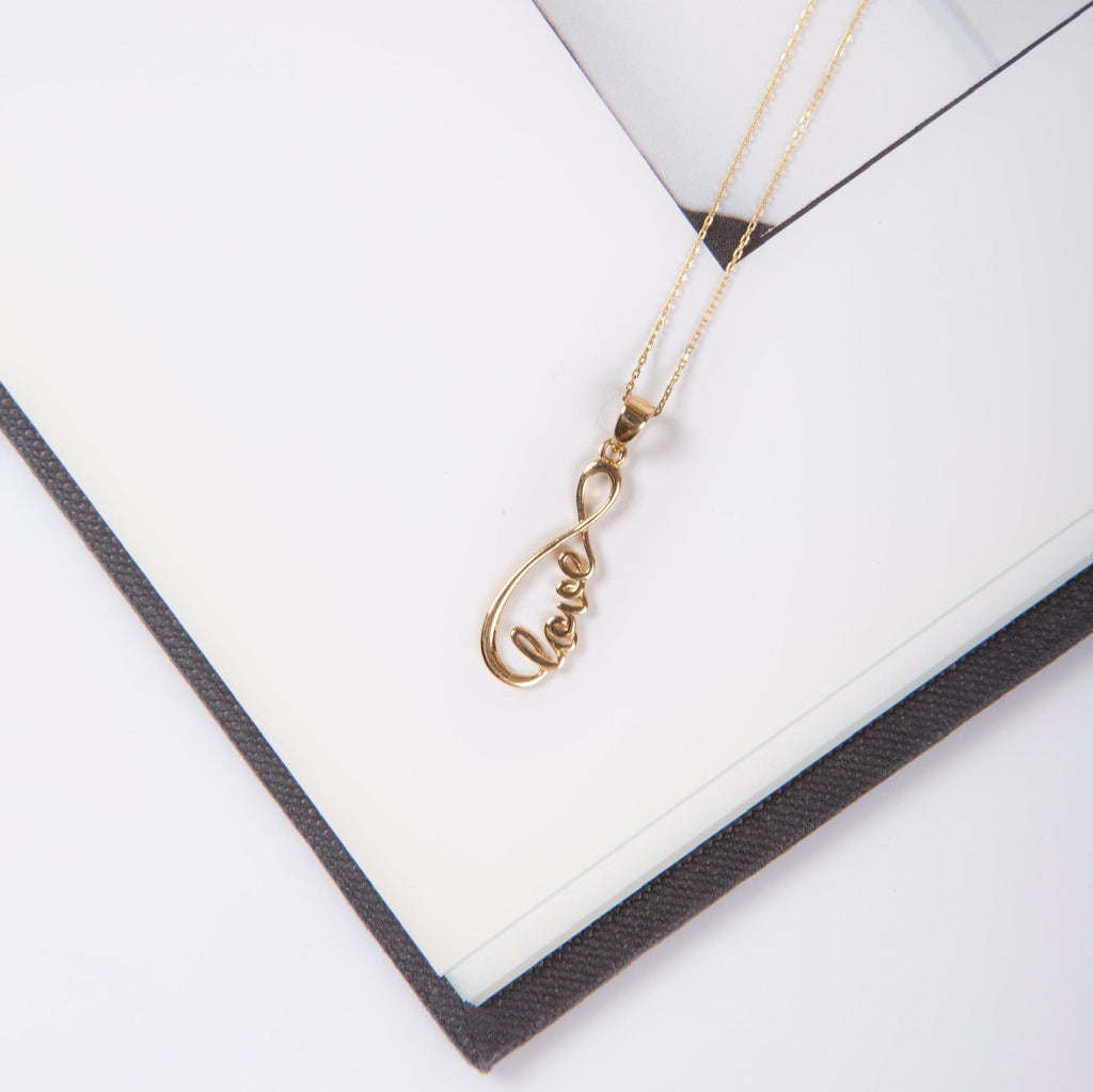 14K Solid Gold Love Infinity Pendant on a chain, elegantly displayed on an open book, tilted to the right, highlighting its delicate infinity and love design.