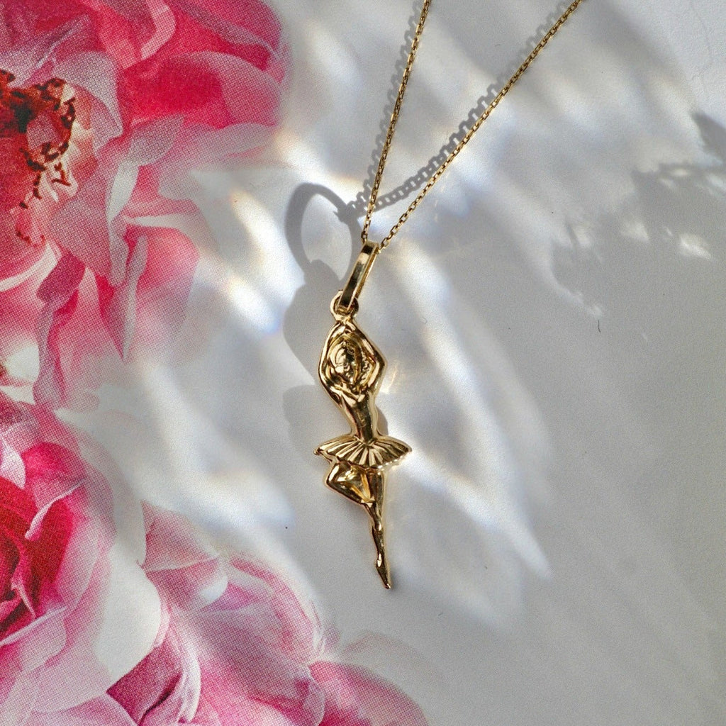 14K Solid Gold Ballerina Charm attached to a chain, laid flat on a smooth surface to showcase its intricate details and polished yellow gold finish.